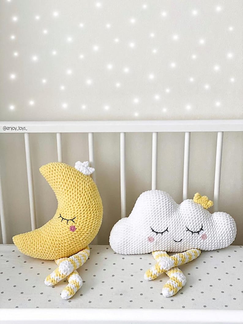 May include: Crocheted nursery decor featuring a yellow crescent moon and a white cloud, both with closed eyes and small floral accents. The moon and cloud have striped yellow and white legs. The items are placed on a crib.