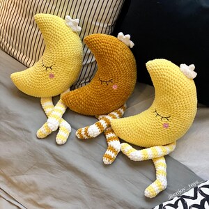 May include: Three crocheted moon-shaped plush toys in yellow and gold, each with closed eyes, rosy cheeks, and a white star-shaped crown. The toys have striped yellow and white legs. The background includes pillows and a bedspread.