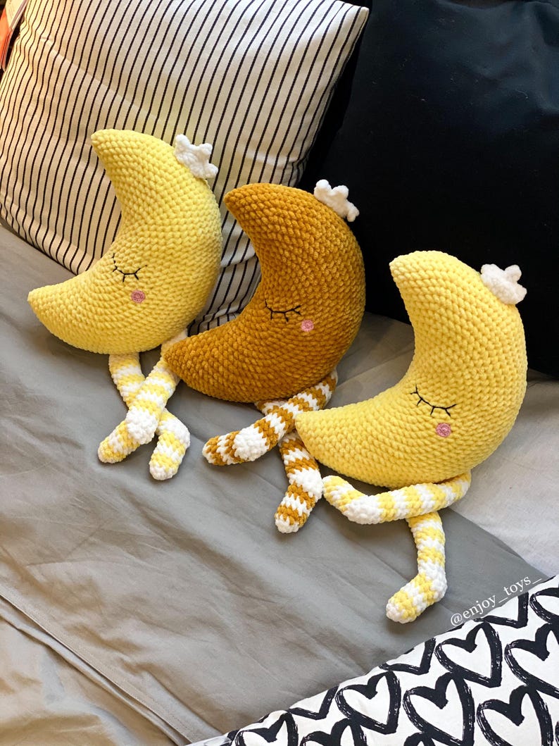 May include: Three crocheted moon-shaped plush toys in yellow and gold, each with closed eyes, rosy cheeks, and a white star-shaped crown. The toys have striped yellow and white legs. The background includes pillows and a bedspread.