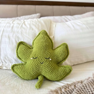 May include: A green, crocheted leaf-shaped cushion with a smiling face and closed eyes. The cushion is a soft, textured item, likely for decorative use. The cushion is resting on a white bed.