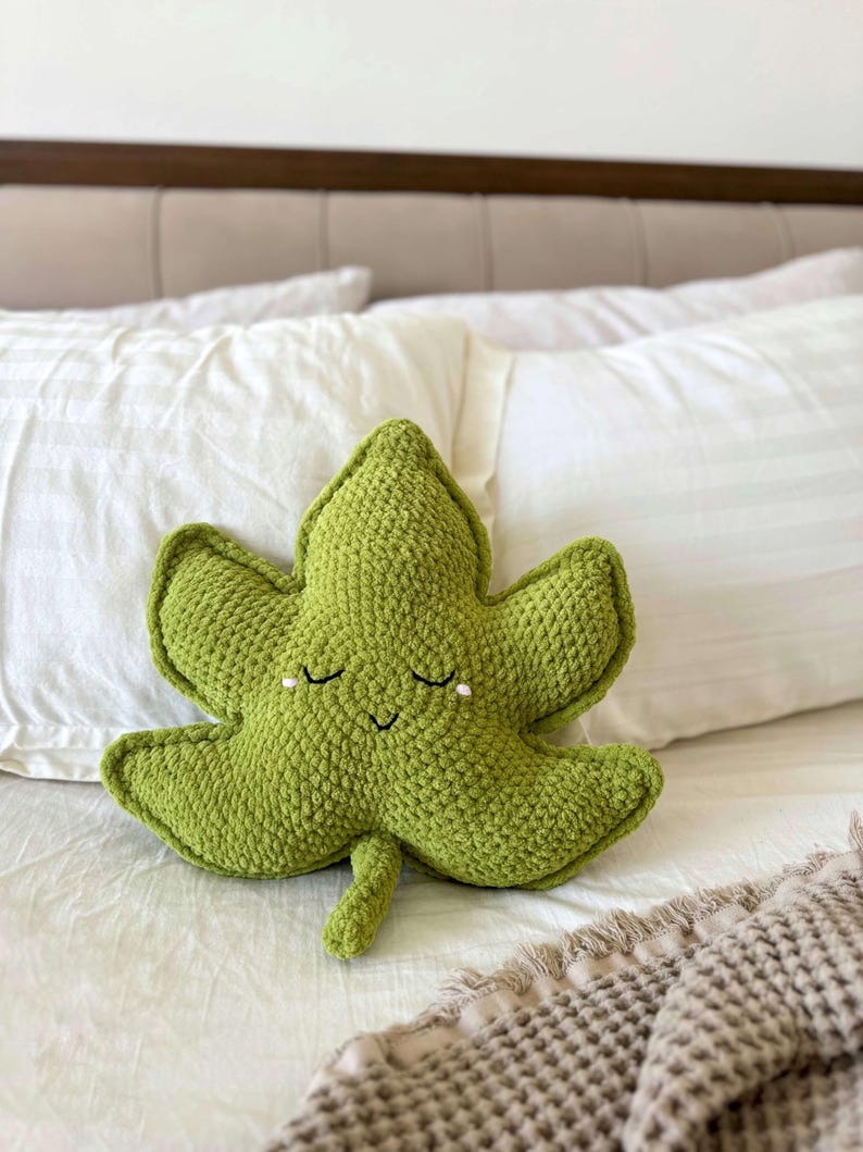 May include: A green, crocheted leaf-shaped cushion with a smiling face and closed eyes. The cushion is a soft, textured item, likely for decorative use. The cushion is resting on a white bed.