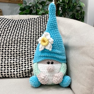 May include: A handmade, plush gnome doll with a light blue pointed hat and a white and yellow flower accent. The gnome has a white beard, a light green and pink face, and blue feet. The doll is sitting on a beige sofa.