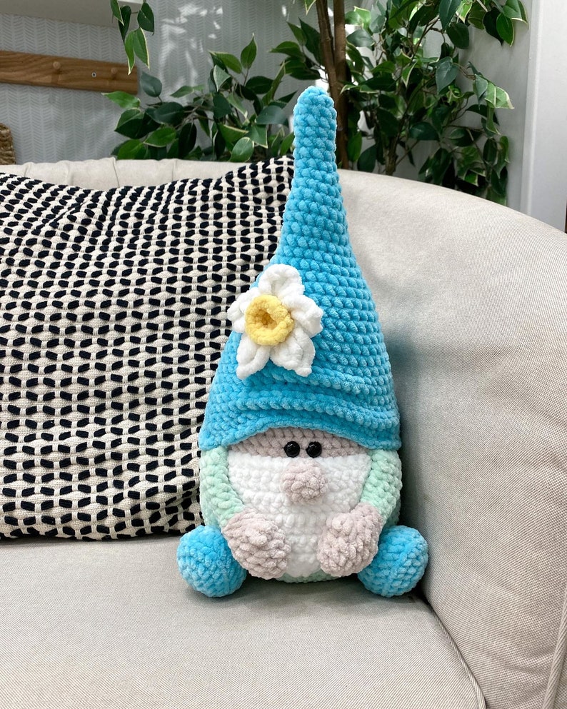 May include: A handmade, plush gnome doll with a light blue pointed hat and a white and yellow flower accent. The gnome has a white beard, a light green and pink face, and blue feet. The doll is sitting on a beige sofa.