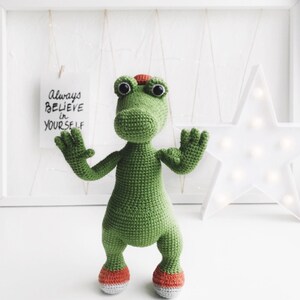 Puede incluir: Juguete de cocodrilo de crochet verde con zapatos naranjas y grandes ojos negros. El juguete est&aacute; de pie sobre una superficie blanca con un cartel que dice "Siempre cree en ti mismo".