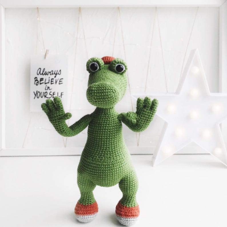 Puede incluir: Juguete de cocodrilo de crochet verde con zapatos naranjas y grandes ojos negros. El juguete est&aacute; de pie sobre una superficie blanca con un cartel que dice "Siempre cree en ti mismo".