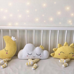May include: Handmade crochet nursery decor featuring a yellow crescent moon, a white cloud, and a yellow sun. Each plush figure has closed eyes and striped legs, resting on a white surface. The background includes a white crib and soft lighting.