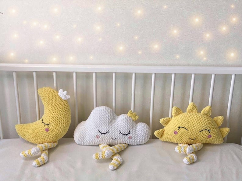 May include: Handmade crochet nursery decor featuring a yellow crescent moon, a white cloud, and a yellow sun. Each plush figure has closed eyes and striped legs, resting on a white surface. The background includes a white crib and soft lighting.