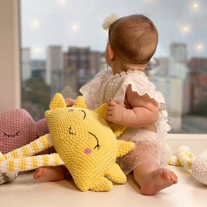 May include: A baby sits on a white surface with three crocheted stuffed toys. The toys are a yellow sun, a pink cloud, and a white cloud. The baby is wearing a white dress with a flower headband.