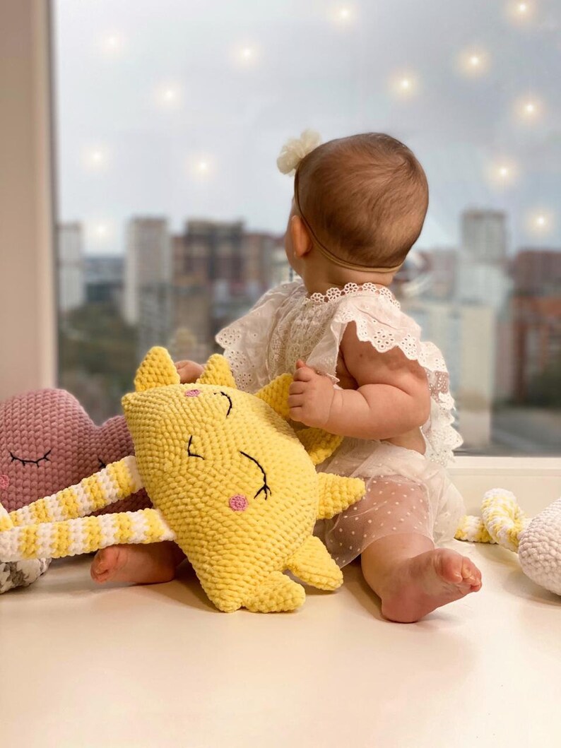 May include: A baby sits on a white surface with three crocheted stuffed toys. The toys are a yellow sun, a pink cloud, and a white cloud. The baby is wearing a white dress with a flower headband.