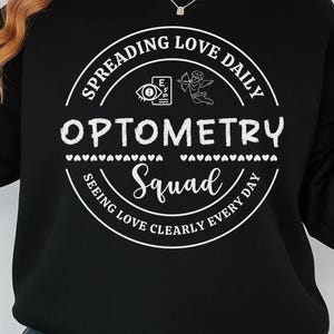 Może przedstawiać: Czarna bluza z okrągłym dekoltem z białym okrągłym nadrukiem. Grafika głosi "Spreading Love Daily Optometry Squad Seeing Love Clearly Every Day". Projekt zawiera tablicę okulistyczną, różę i elementy dekoracyjne.