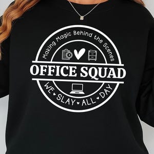 May include: Black sweatshirt with a white graphic design that says "Making Magic Behind the Scenes Office Squad We Slay All Day". The graphic includes a heart, a camera, a file folder, and a desk.