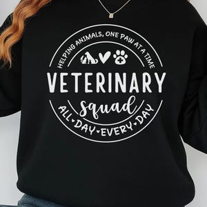 May include: Black sweatshirt with white text that reads "VETERINARY squad ALL DAY EVERY DAY" inside a circular design. The design includes paw prints and animal silhouettes. The phrase "Helping animals, one paw at a time" is also included.