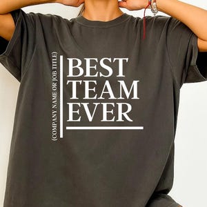 Peut inclure: T-shirt gris foncé avec l'inscription blanche "BEST TEAM EVER" et le texte "(COMPANY NAME OR JOB TITLE)" sur le côté gauche. Le t-shirt a des manches courtes et une coupe ample.