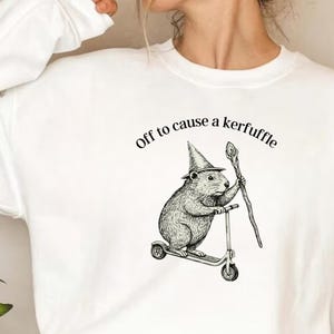 Könnte beinhalten: Weißes Sweatshirt mit einer Grafik eines Nagetiers mit Hexenhut, das einen Roller fährt und einen Stab hält. Der Text "off to cause a kerfuffle" ist über der Illustration angeordnet.