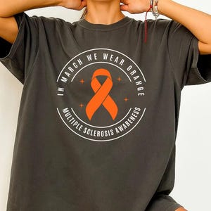 Op de afbeelding: Donkergrijs t-shirt met een cirkelvormig ontwerp met een oranje lint en de tekst "IN MARCH WE WEAR ORANGE MULTIPLE SCLEROSIS AWARENESS". Het shirt heeft korte mouwen en een relaxte pasvorm.
