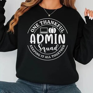 May include: Black sweatshirt with a white circular graphic. The graphic reads "ONE THANKFUL ADMIN Squad KEEPING IT ALL TOGETHER" with a laptop and pumpkin illustration. The sweatshirt has long sleeves and a crew neck.