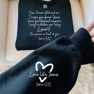 May include: Black sweatshirt with white text that reads "Dear Person Behind me, I hope you know Jesus loves you beyond measure I pray he blesses you today Love -The person in front of you John 15:12" and a heart. The bottom of the sweatshirt has a white heart with the text "Love like Jesus" and "John 15:12".