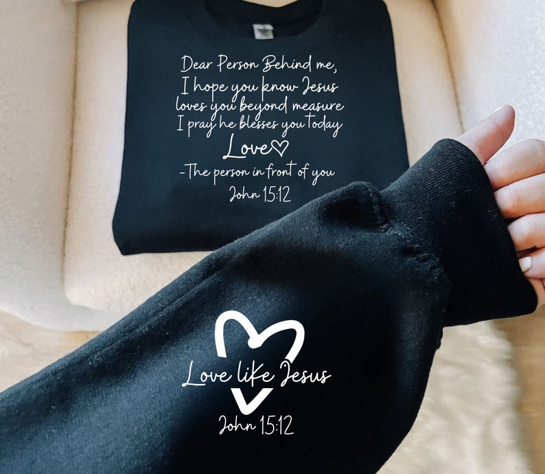 May include: Black sweatshirt with white text that reads "Dear Person Behind me, I hope you know Jesus loves you beyond measure I pray he blesses you today Love -The person in front of you John 15:12" and a heart. The bottom of the sweatshirt has a white heart with the text "Love like Jesus" and "John 15:12".