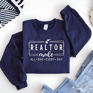 May include: Navy blue sweatshirt with white text that reads "Realtor mode - All Day Every Day" with a house and key graphic.