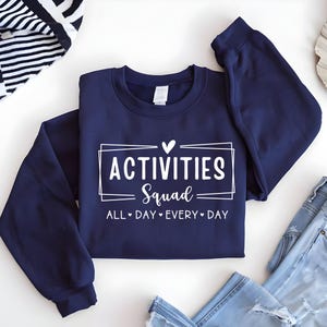 Op de afbeelding: Navyblauwe trui met witte tekst die "Activities Squad All Day Every Day" met een hartje zegt.