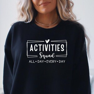 Op de afbeelding: Zwart sweatshirt met witte tekst die "Activities Squad All Day Every Day" luidt met een hartje tussen elk woord.