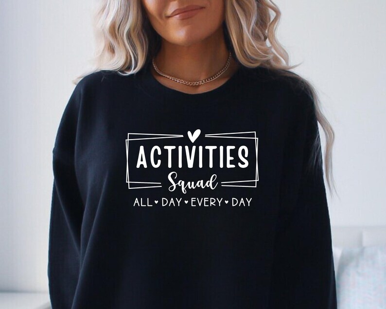 Op de afbeelding: Zwart sweatshirt met witte tekst die "Activities Squad All Day Every Day" luidt met een hartje tussen elk woord.