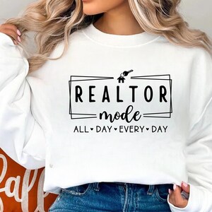 May include: White sweatshirt with black text that reads "Realtor Mode All Day Every Day" with a house and key graphic.