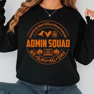 May include: Black sweatshirt with an orange graphic that reads "ADMIN SQUAD" in a circular design with Halloween-themed icons and the phrases "Haunting the Office Since Forever" and "We Slay All Day."