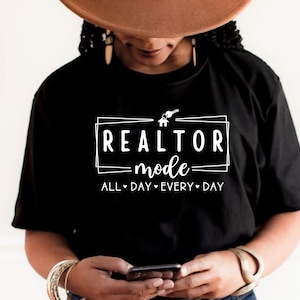 May include: Black t-shirt with white text that reads "Realtor Mode" in a box with a house and key icon. The text below the box reads "All Day Every Day".