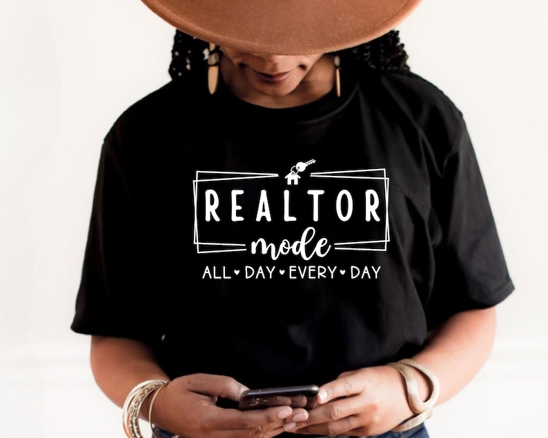 May include: Black t-shirt with white text that reads "Realtor Mode" in a box with a house and key icon. The text below the box reads "All Day Every Day".