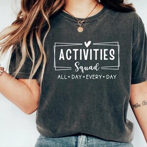 Op de afbeelding: Een donkergrijs T-shirt met een witte afbeelding die "Activities Squad" zegt met een hart boven het woord "Squad" en de tekst "All Day Every Day" eronder.