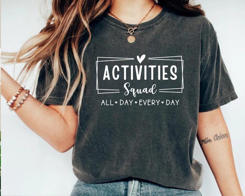 Op de afbeelding: Een donkergrijs T-shirt met een witte afbeelding die "Activities Squad" zegt met een hart boven het woord "Squad" en de tekst "All Day Every Day" eronder.