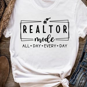 May include: White t-shirt with black text that reads "Realtor mode all day every day" with a house and key graphic above the text.