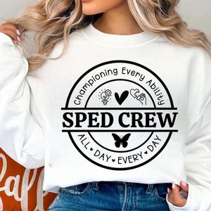 Puede incluir: Sudadera blanca con un diseño circular negro que dice "Championing Every Ability" y "SPED CREW" con una bombilla, un corazón y manos sosteniendo un corazón. El círculo también dice "All Day Every Day" con una mariposa.