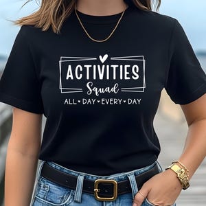 Op de afbeelding: Zwart T-shirt met witte tekst die "Activities Squad" zegt met een hartje en "All Day Every Day" eronder.