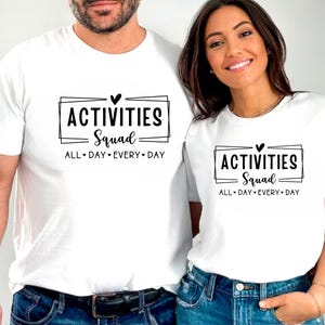 Op de afbeelding: Twee witte T-shirts met zwarte tekst die "Activities Squad All Day Every Day" zegt en een kleine hartgrafiek.