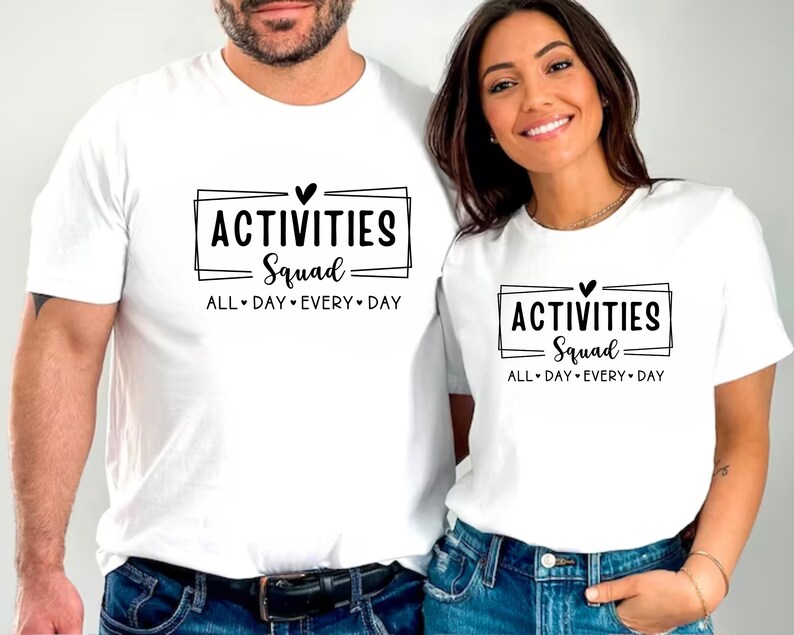 Op de afbeelding: Twee witte T-shirts met zwarte tekst die "Activities Squad All Day Every Day" zegt en een kleine hartgrafiek.