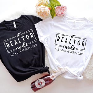 May include: Two t-shirts with the text "REALTOR mode ALL DAY EVERY DAY" in a black box with a house and key graphic. The t-shirt on the left is black and the t-shirt on the right is white.