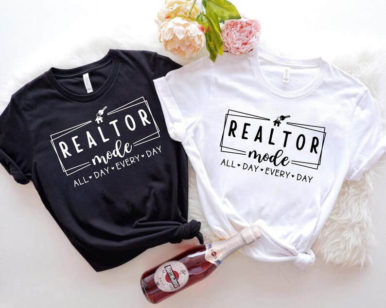 May include: Two t-shirts with the text "REALTOR mode ALL DAY EVERY DAY" in a black box with a house and key graphic. The t-shirt on the left is black and the t-shirt on the right is white.