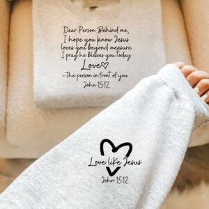 May include: Gray sweatshirt with a black heart and the text "Love like Jesus" and "John 15:12". The text "Dear Person Behind me, I hope you know Jesus loves you beyond measure. I pray he blesses you today. Love - The person in front of you John 15:12" is visible on a white towel.