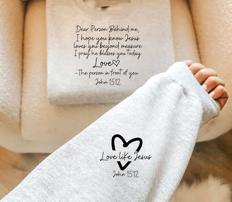 May include: Gray sweatshirt with a black heart and the text "Love like Jesus" and "John 15:12". The text "Dear Person Behind me, I hope you know Jesus loves you beyond measure. I pray he blesses you today. Love - The person in front of you John 15:12" is visible on a white towel.