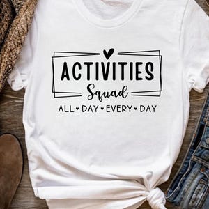Op de afbeelding: Wit T-shirt met zwarte tekst die "Activities Squad All Day Every Day" luidt met een hartje tussen elk woord.