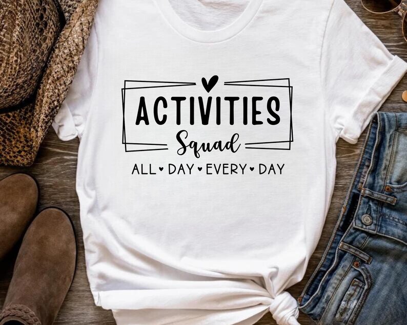 Op de afbeelding: Wit T-shirt met zwarte tekst die "Activities Squad All Day Every Day" luidt met een hartje tussen elk woord.