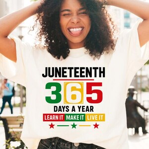 May include: White t-shirt with the text "Juneteenth 365 Days a Year Learn It Make It Live It" in black and green, red, and yellow lettering. There are three red stars below the text.