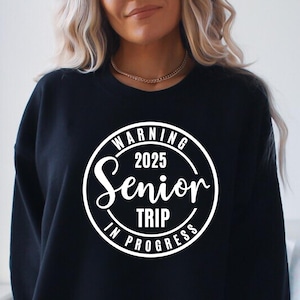 Puede incluir: Sudadera negra con un gráfico circular blanco que dice "Warning 2025 Senior Trip In Progress".