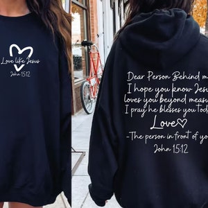 May include: Two black sweatshirts with white text. The front sweatshirt has a heart and the text "Love like Jesus John 15:12". The back sweatshirt has the text "Dear Person Behind me, I hope you know Jesus loves you beyond measure I pray he blesses you today Love♡ -The person in front of you John 15:12".