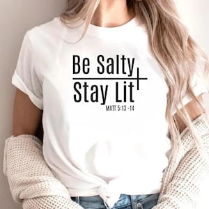 以下が含まれることがあります： 黒い文字で「Be Salty Stay Lit」と書かれた白いTシャツ。下に聖書の引用「MATT 5:13-14」が書かれている。