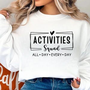 Op de afbeelding: Witte trui met zwarte tekst die "Activities Squad" met een hartje en "All Day Every Day" eronder staat.