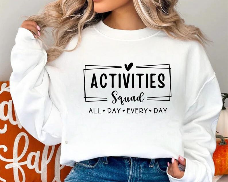 Op de afbeelding: Witte trui met zwarte tekst die "Activities Squad" met een hartje en "All Day Every Day" eronder staat.