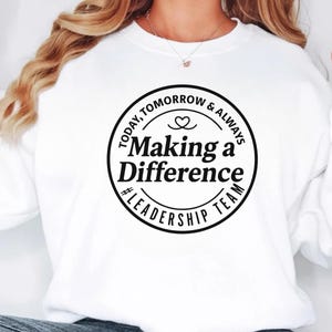 Pode incluir: Camisola branca com um design circular preto que diz "Today, Tomorrow & Always Making a Difference #Leadership Team" com um símbolo de coração. A camisola é de estilo casual.